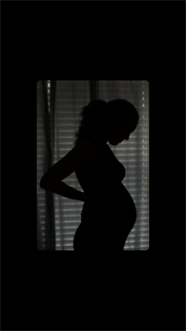 Silhouette of a pregnant person standing in front of window blinds